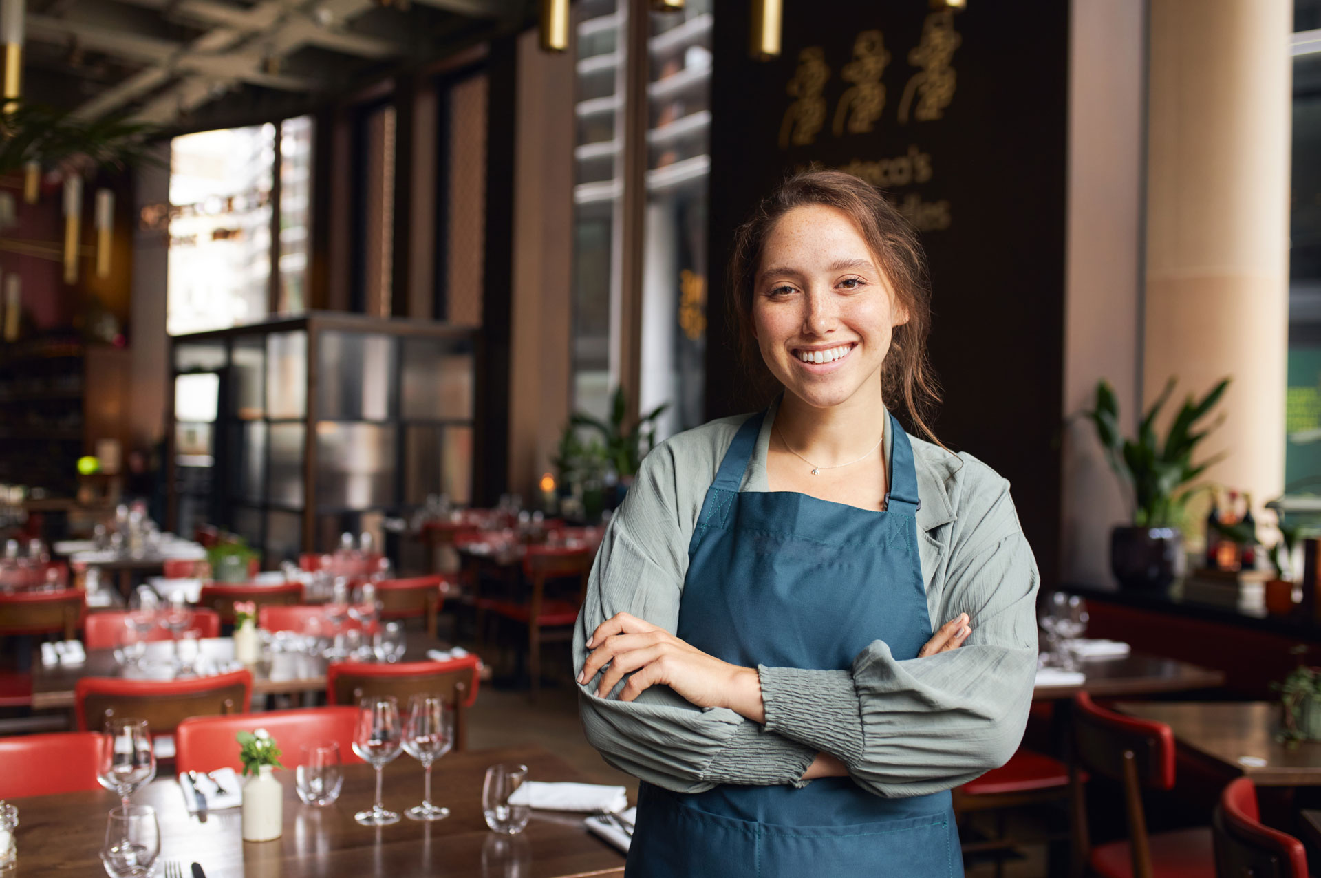 Lächelnde Mitarbeiterin mit Schürze steht mit verschränkten Armen in einem Restaurant